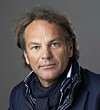 Portrait of Per G Braathen wearing a dark jacket and scarf against a grey background.