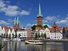 Byhorisont med røde tage, kirketårne, brostenslignende bygninger og en båd på vandet under blå himmel.