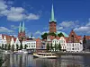 Byhorisont med røde tage, kirketårne, brostenslignende bygninger og en båd på vandet under blå himmel.