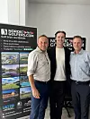 Tre mænd står indendørs foran to NordicGolfers.com-rollups.