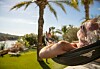 En person ligger i en hængekøje ved en pool med palmer og udsigt til vandet i baggrunden.
