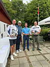 Fire mænd står på en terrasse og holder præmier ved en golfklub med et hvidt telt og grønne træer i baggrunden.