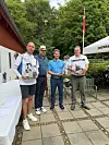 Fire mænd står på en terrasse og holder præmier ved en golfklub med et hvidt telt og grønne træer i baggrunden.