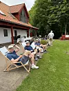 Flere personer sidder i havestole på en terrasse ved et klubhus med rød tag og grønne omgivelser.