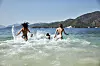Tre personer springer och badar i vågorna vid en kuststrand med berg och klart blått vatten.