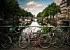 Cyklar står parkerade vid en kanal med båtar och träd längs vattnet i Amsterdam.