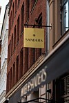 Gult SANDERS-skilt hænger ud fra en rød murstensbygning over en butiksskilting.
