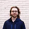Nuori mies, jolla on silmälasit ja tummansininen takki, seisoo vaaleaa tiiliseinää vasten.