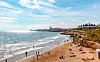 Bred sandstrand i Sitges i Spanien med människor som badar och solar, havet till vänster och bebyggelse på klippor i fjärran.