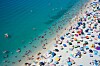 Flera rader av färgglada parasoller och människor på en fullsatt strand vid turkost vatten.