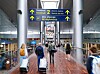 Resenärer med rullväskor går genom en flygplatsterminal med skyltar mot avgångar och incheckning.