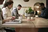Flera personer arbetar med laptops vid ett träbord med kaffe och en växt i en hotellounge.